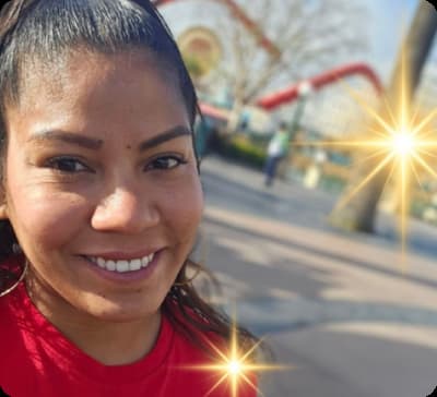 A smiling woman is wearing a red shirt, with her hair tied back. She is outdoors, with a blurred background featuring red structures and trees, enhanced by glowing light effects.