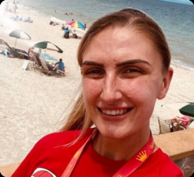 a person smiling at the beach. The background includes sand, umbrellas, and people relaxing by the sea. The individual is wearing a red shirt with a visible lanyard