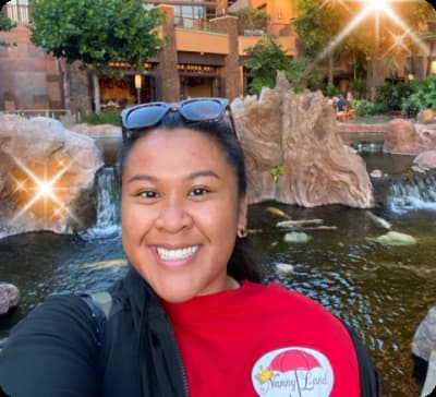 A smiling woman is wearing a red shirt with a "Nanny Land" logo, standing in front of a pond with rocks, waterfalls, and greenery, with a glowing light effect in the background